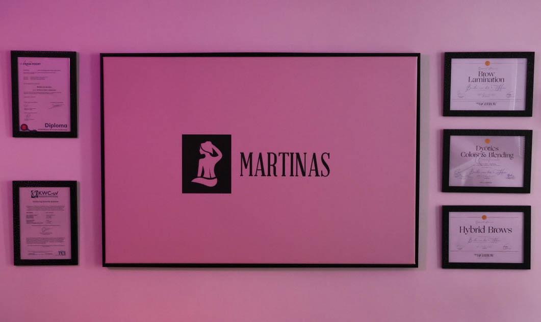 De roze salonwand met het ingelijste Martinas-logo en vier diploma's